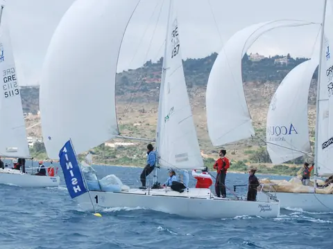 Three J/24 keelboats running under white spinnakers in a strong breeze, his boat in the centre, number 1 on the bow, crossing the finish beside a blue FINISH flag ahead of the other two